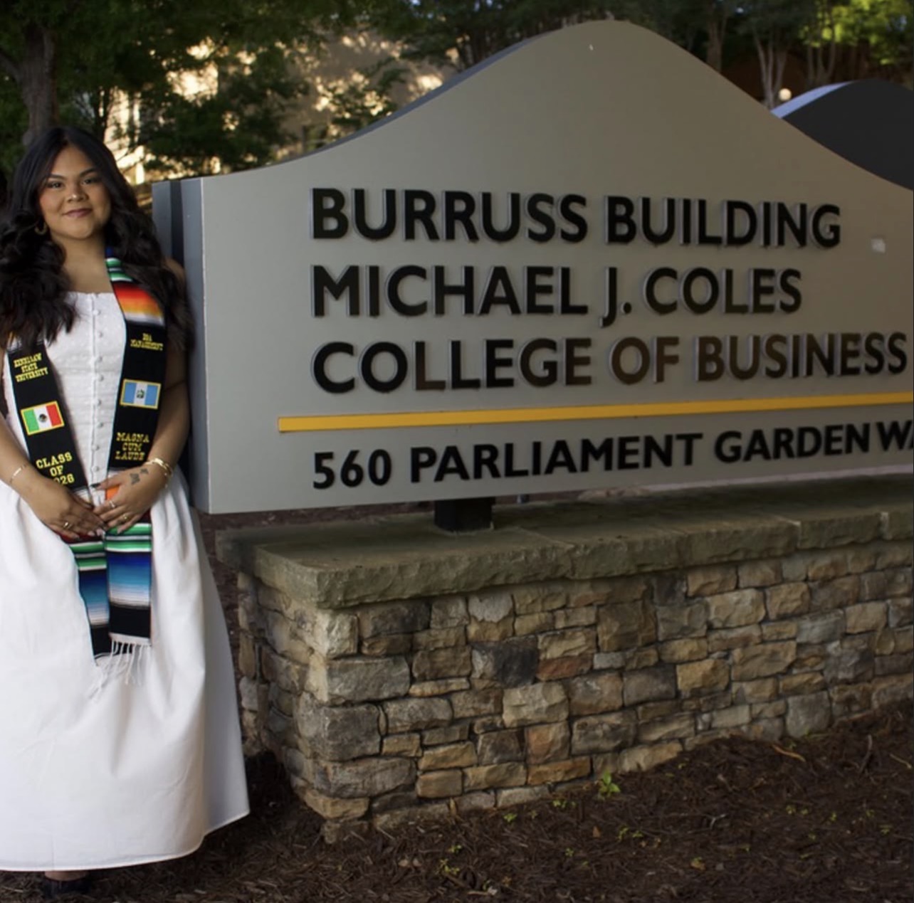 Graduate posing beside the Michael J. Coles College of Business sign